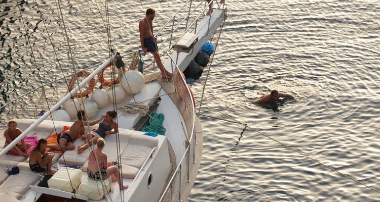 People relaxing on a sailboat with one person swimming in the water.