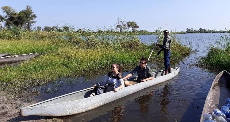 Zwei Personen werden in einem kleinen Boot durch einen Fluss oder Kanal mit grasbewachsenen Ufern geführt.