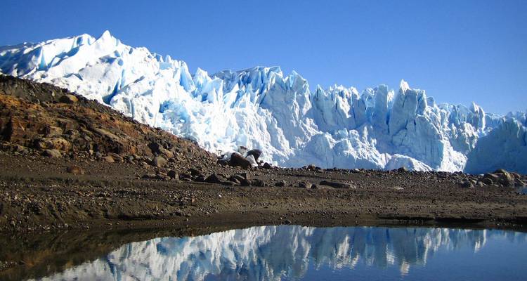 Aufragende Eismauer aus blauem Eis des Perito-Moreno-Gletschers, die sich in einer stillen Lagune unter klarem Himmel spiegelt.