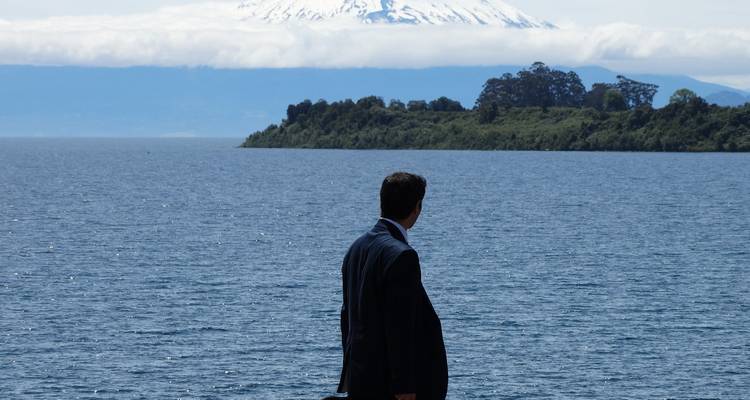 Una persona vestida con traje mirando un lago con montañas nevadas de fondo.