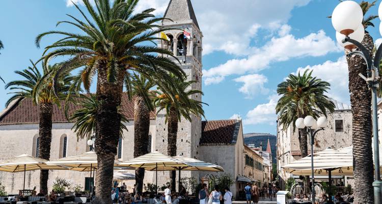 Place animée ombragée par les palmiers à Trogir avec des cafés en plein air et le clocher qui s'élève à l'arrière.
