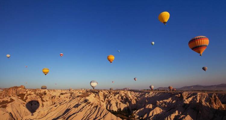 Docenas de globos aerostáticos coloridos flotan sobre los valles ondulados de arenisca de Capadocia al amanecer bajo un cielo azul despejado.