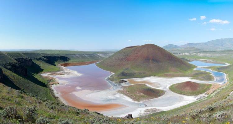 Vista panorámica de un cráter volcánico de bordes rojos rodeado de lagos coloridos poco profundos y llanuras verdes bajo un cielo despejado.