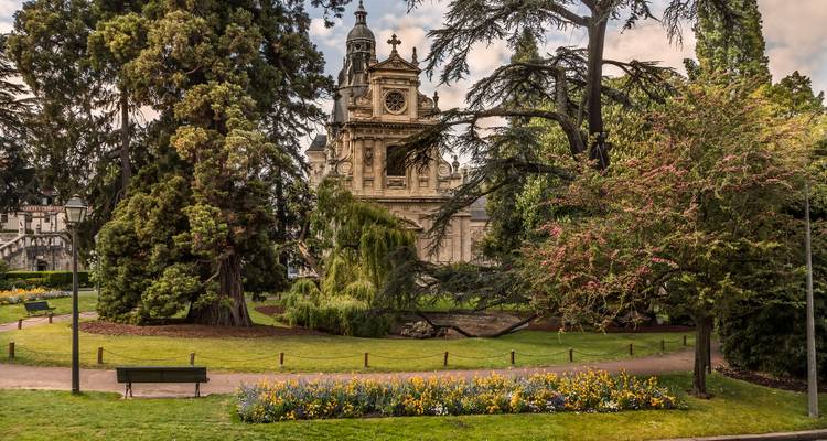 Schaduwrijke stadspark met bloeiende bloemperken die een prachtige barokke kerkgevel omlijsten achter volwassen bomen.