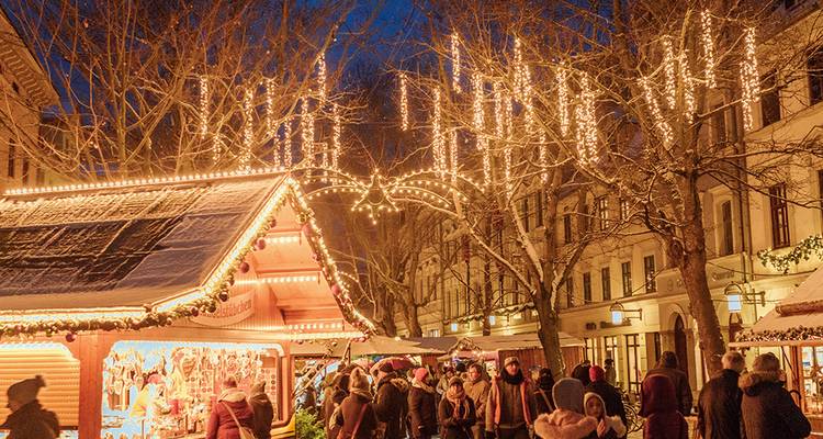 Mercado navideño festivo al atardecer con luces centelleantes, puestos de madera y visitantes abrigados.
