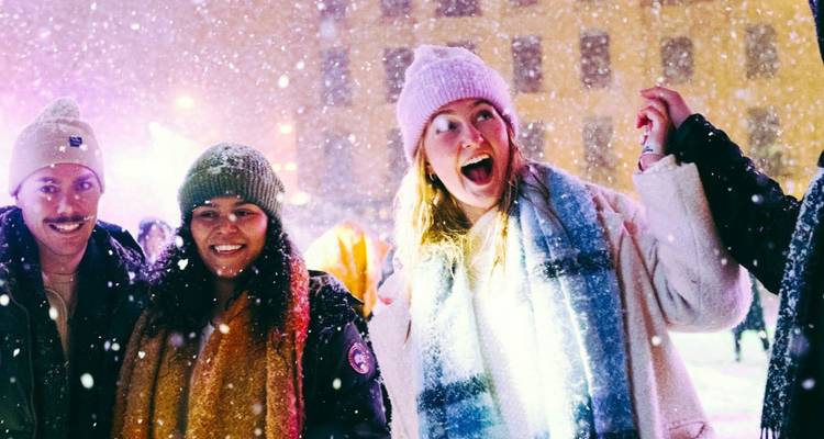 Fröhliche Freunde in Wintermänteln und Mützen lachen zusammen, während Schnee auf einem Stadtplatz fällt, der von farbigen Lichtern beleuchtet wird.