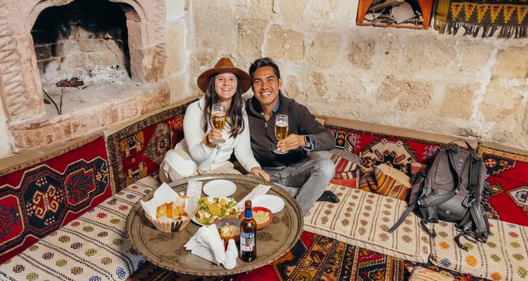 Pareja sonriente disfrutando bebidas y meze turco en asientos acolchados en un restaurante con paredes de piedra