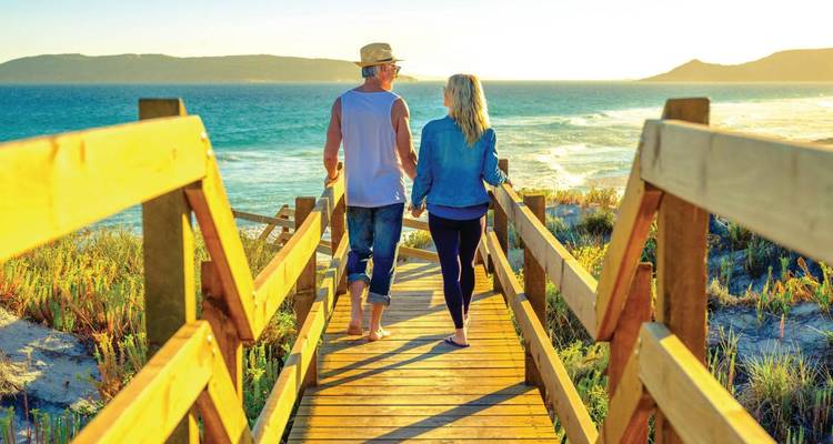 Couple marchant pieds nus sur une passerelle en bois en direction d'un océan turquoise au coucher du soleil