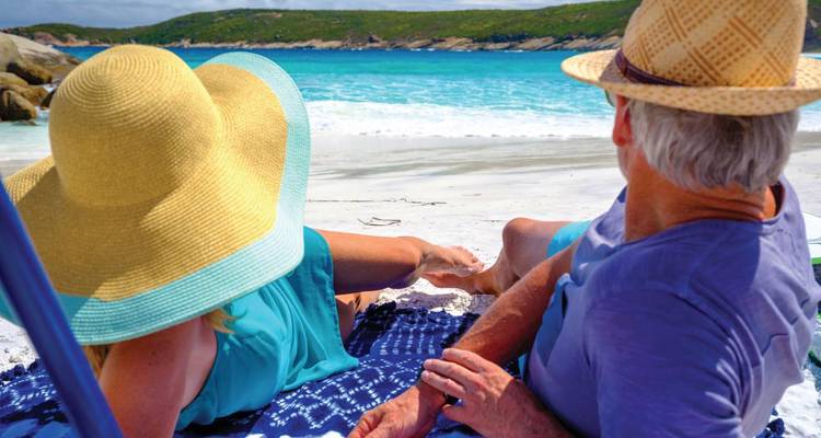 Couple de personnes âgées se détendant sur une plage de sable blanc avec une eau turquoise, portant de larges chapeaux de soleil