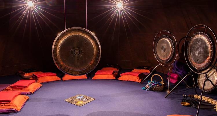 Salle de méditation circulaire faiblement éclairée avec gongs, coussins et moquette violet doux.