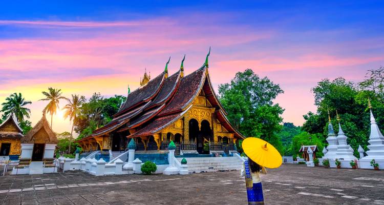 Vibrante atardecer sobre Wat Xieng Thong en Luang Prabang con una mujer vestida con túnica sosteniendo una sombrilla amarilla.