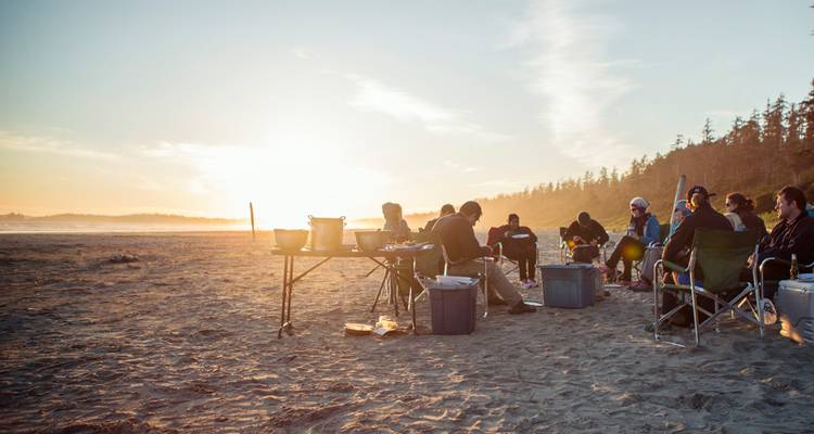 Un grand groupe profite d'un repas en plein air sur la plage au coucher du soleil avec des chaises de camping et des tables installées sur le sable.