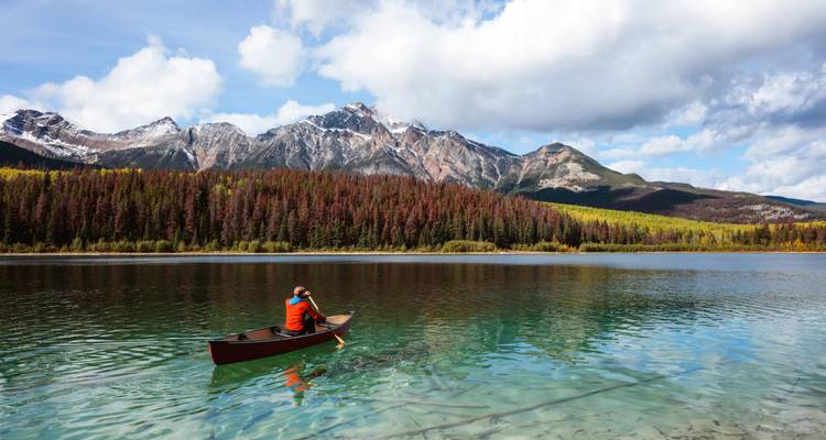 Un pagayeur dans un canoë rouge glisse sur un lac de montagne d'une clarté cristalline qui reflète les pics enneigés et la forêt d'automne.