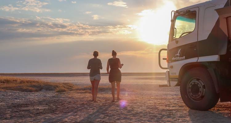 Deux personnes marchant sur un désert de sel près d'un grand camion de tourisme au coucher du soleil.