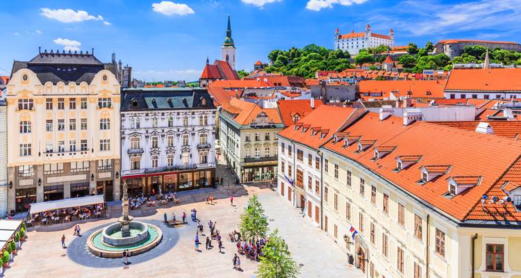 Vista panorámica de la plaza principal de Bratislava con fachadas pastel, fuente central, aguja de la catedral y castillo en la colina.
