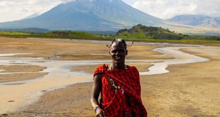 Homme de la tribu Masaï en shuka rouge debout dans un lit de rivière aride avec un paysage de montagne volcanique en arrière-plan