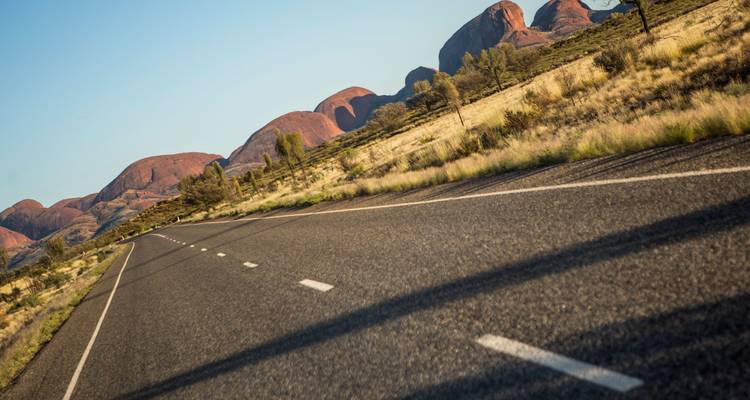 Empty highway curves toward the domed orange rocks of Kata Tjuta beneath bright blue skies.