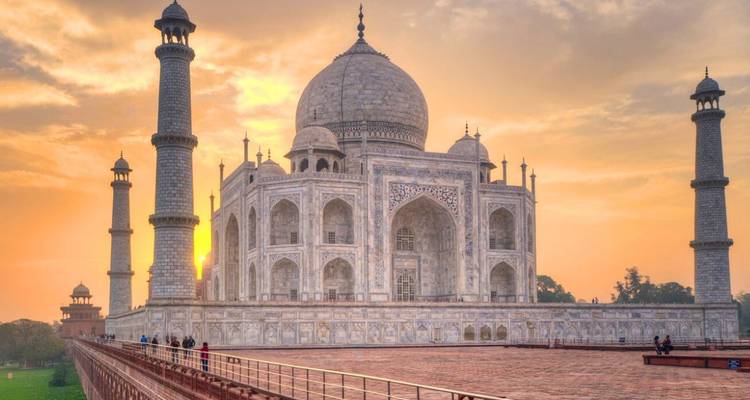 Taj Mahal glowing at sunrise with golden sky and visitors on red sandstone platform.