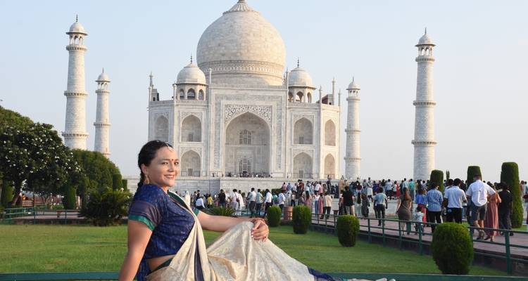 Woman in blue sari posing on bench with bustling Taj Mahal grounds behind.