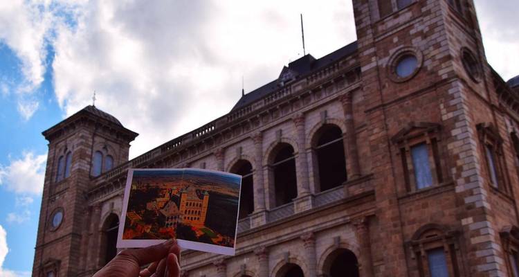 Une carte postale tenue contre la façade d'un palais historique en pierre sous un ciel partiellement nuageux.
