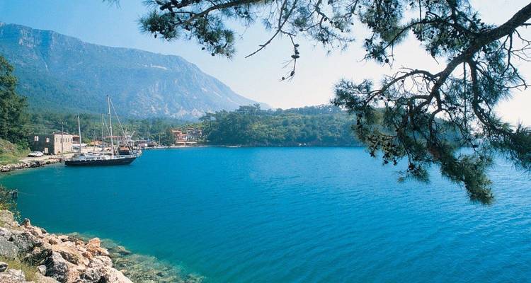 Coastal bay with clear blue water, a sailing ship, and mountainous backdrop.