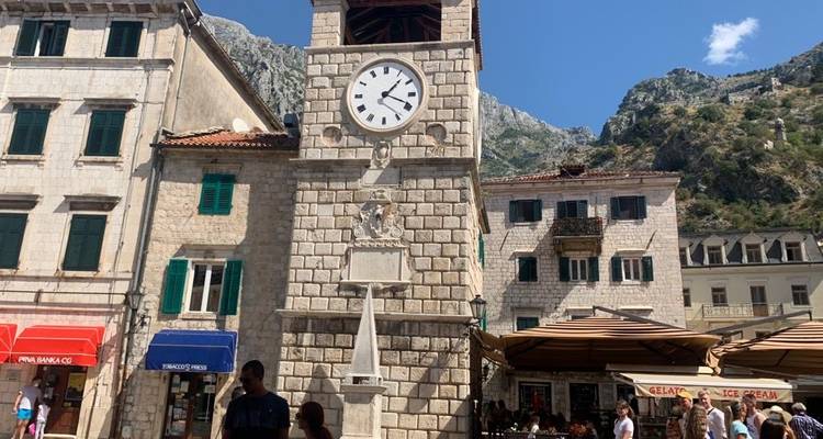 Tour de l'horloge entourée de bâtiments en pierre et d'une place animée.