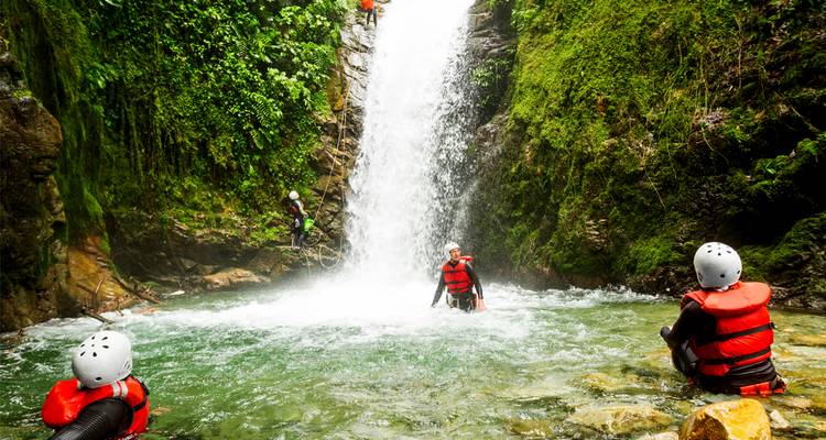 Des aventuriers portant des casques et des gilets de sauvetage pataugent sous une cascade de jungle luxuriante lors d'une séance de canyoning.