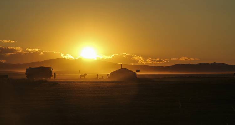 Coucher de soleil sur une plaine herbeuse avec une yourte et une silhouette à cheval au loin.
