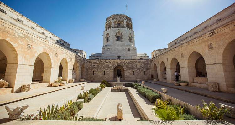 Cour en pierre avec un jardin central en contrebas et une haute tour ronde encadrée par des murs à arcades sous un ciel bleu dégagé.