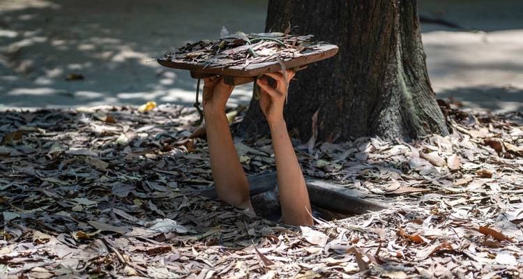 Los brazos emergen de una entrada de túnel camuflada levantando una tapa de hojas junto a un árbol.