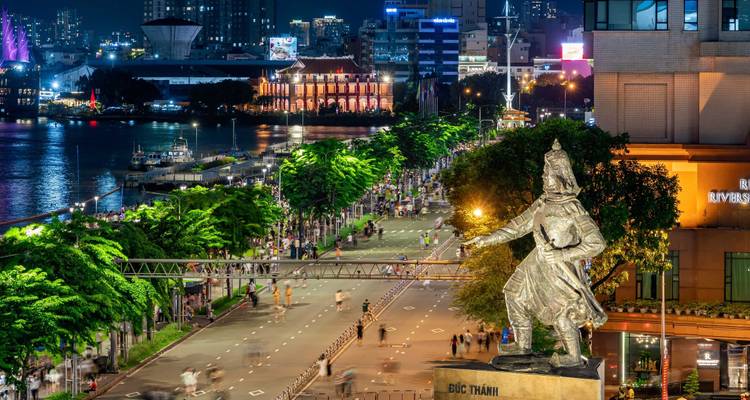 Vista nocturna de un bulevar junto al río con multitudes, luces brillantes y una gran estatua histórica.