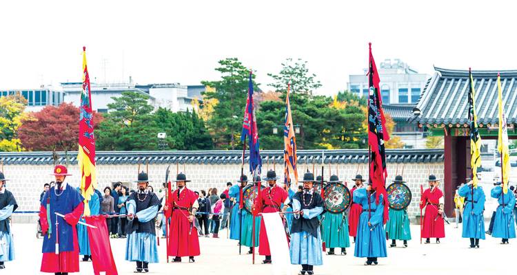 Des gardes en uniformes colorés exécutent une cérémonie royale de relève de la garde devant les grandes portes d'un palais.