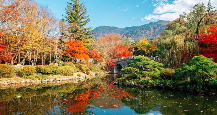 Jardin automnal coloré avec un pont en pierre enjambant un étang reflétissant et des montagnes boisées au-delà