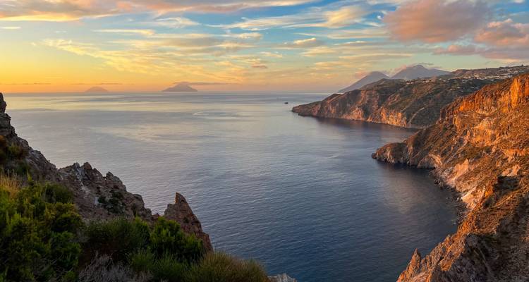 Vue sur l'océan au coucher du soleil avec des îles lointaines et une côte rocheuse.
