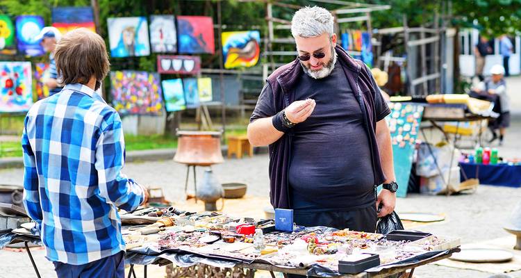 Mannen die items bekijken op een buitenmarkt met kleurrijke kunst op de achtergrond.