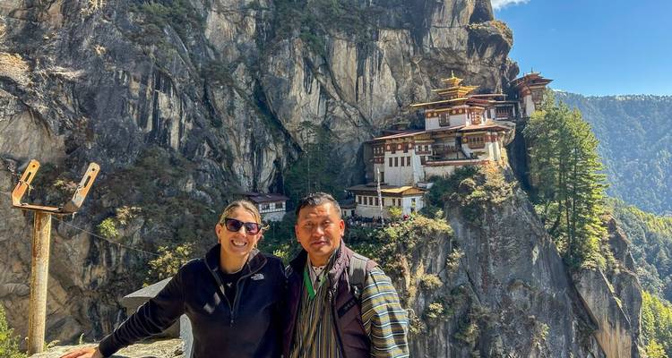 Dos excursionistas se encuentran en el borde del acantilado con el Monasterio del Nido del Tigre de Bután dramáticamente encaramado en la roca detrás de ellos.
