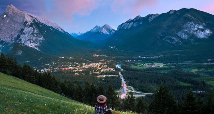 Solo traveler sitting on a grassy slope overlooking Banff town and surrounding Rocky Mountains.