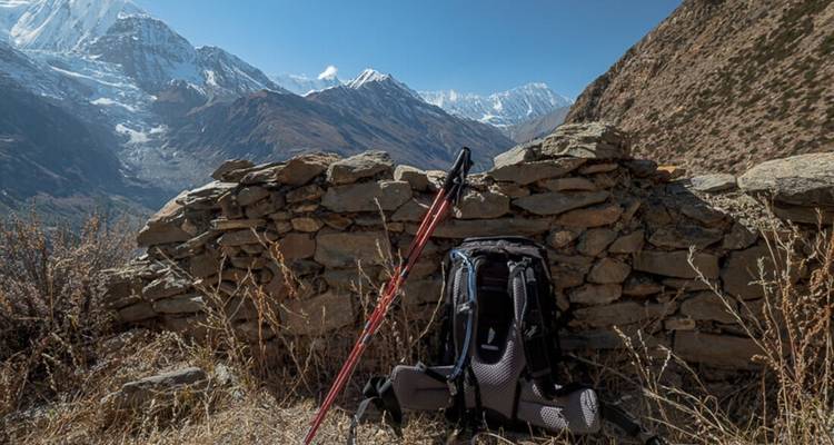 Rucksack und Wanderstöcke lehnen an einer Steinmauer mit Bergen.