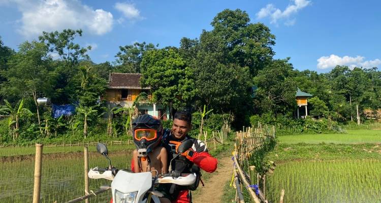 Zwei Personen auf einem Motorrad in einer ländlichen Umgebung mit Häusern und Grünflächen.