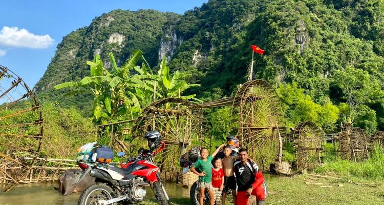 Vier Personen posieren mit einem Motorrad vor Bambuswasserrädern und üppigen Hügeln.