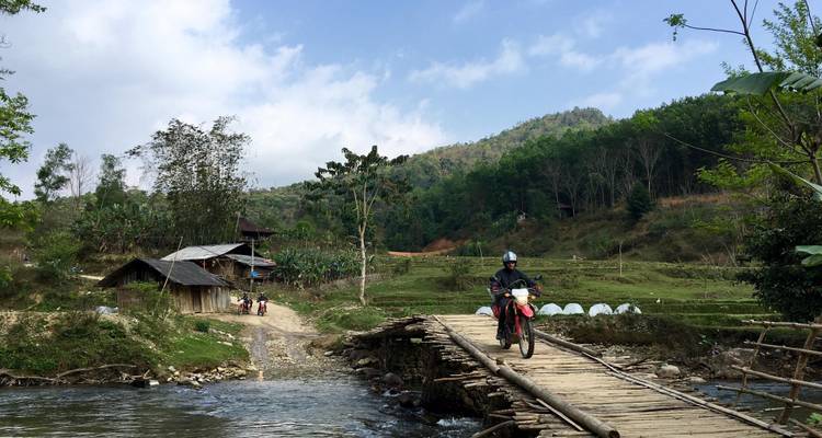 Eine Person auf einem Motorrad überquert eine Holzbrücke über einen Fluss.