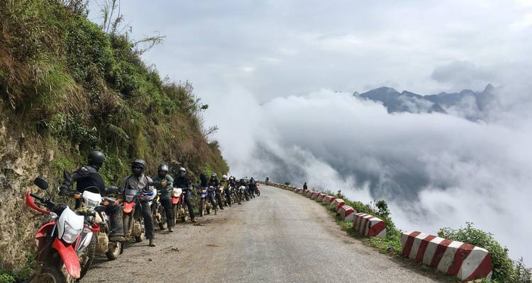 Groep motorrijders op een weg boven de wolken met bergtoppen in de verte.