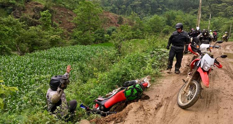 Man naast een omgevallen motorfiets op een modderig pad met een andere man die toekijkt.