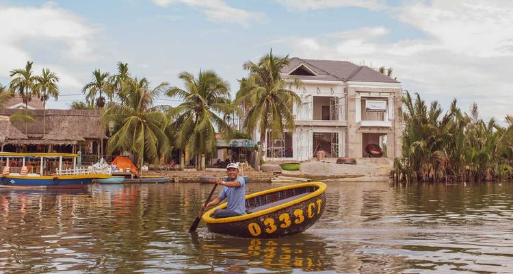 Man paddling a circular boat in front of a building on the riverbank.