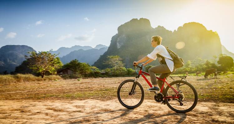 Person cycling on a dirt path with mountains and setting sun in the background.