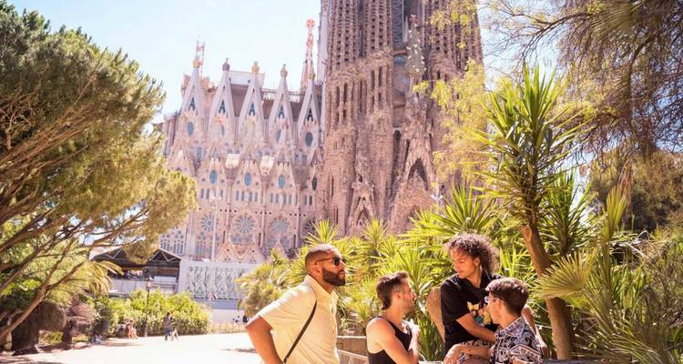 Groupe d'amis discutant et souriant dans un parc bordé de palmiers avec l'emblématique Sagrada Familia qui s'élève derrière eux.