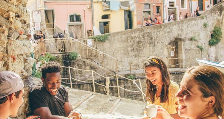Cuatro viajeros disfrutan de café y conversación en una terraza soleada con vista al pintoresco sendero del puerto de Cinque Terre