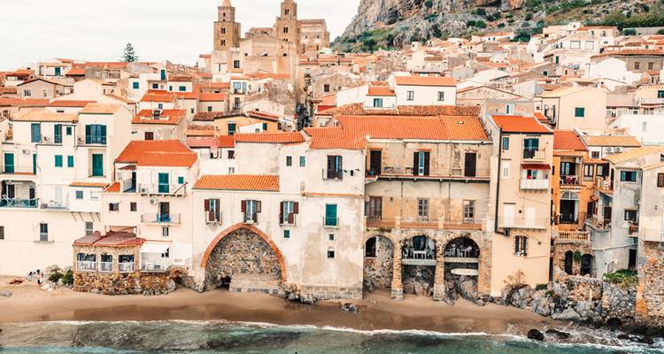 Histórica ciudad costera de Cefalù con arcos de piedra, playa de arena y iglesias medievales bajo una colina rocosa.