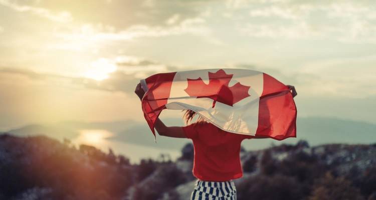 Un voyageur à contre-jour agite un drapeau canadien devant un coucher de soleil montagneux voilé par la brume.