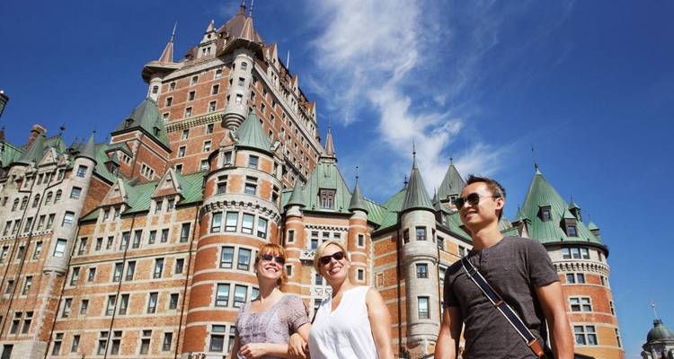 Trois touristes souriants marchent sous le majestueux Château Frontenac dans un ciel bleu éclatant.